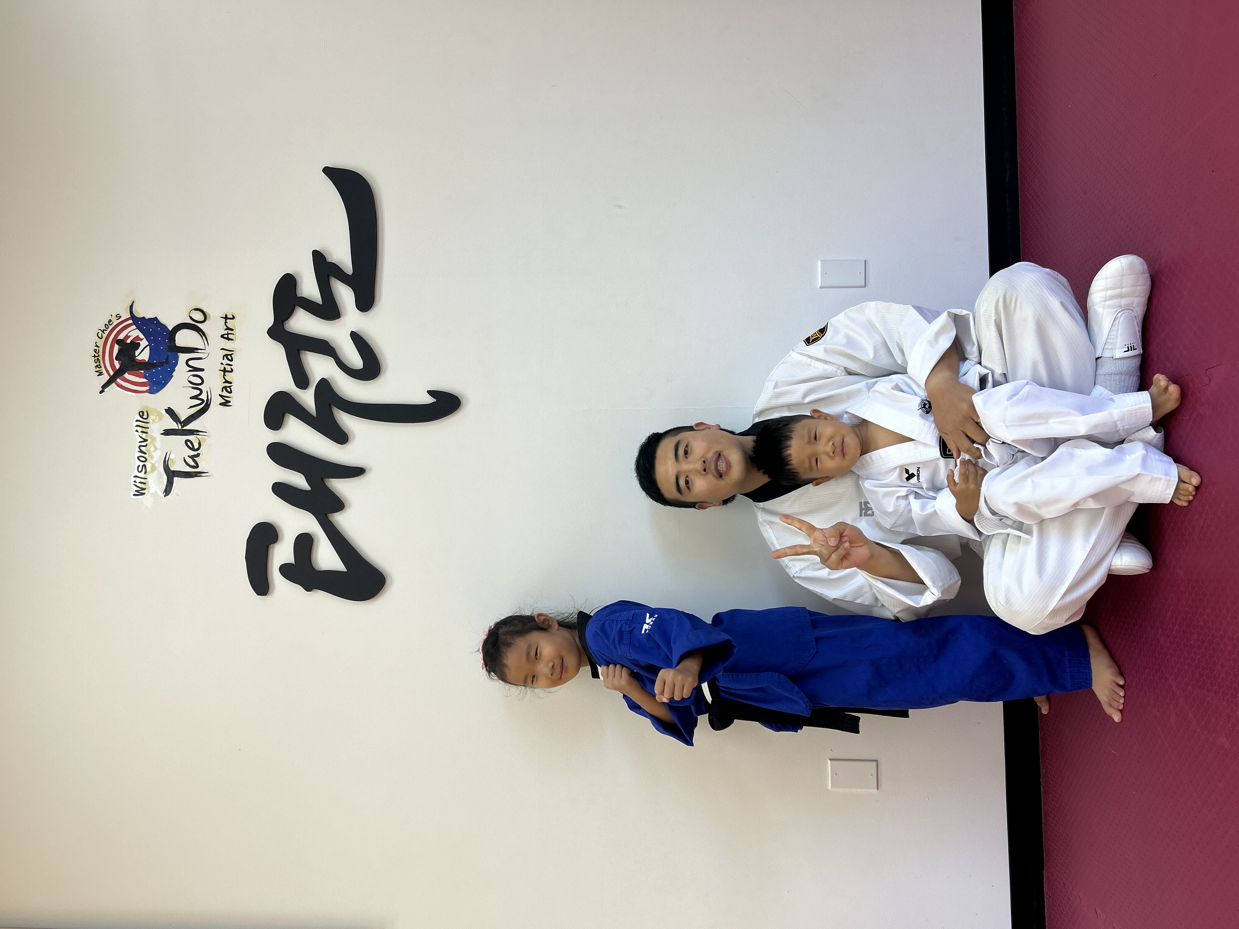 Instructor sitting with two young students beneath the academy logo and Taekwondo calligraphy
